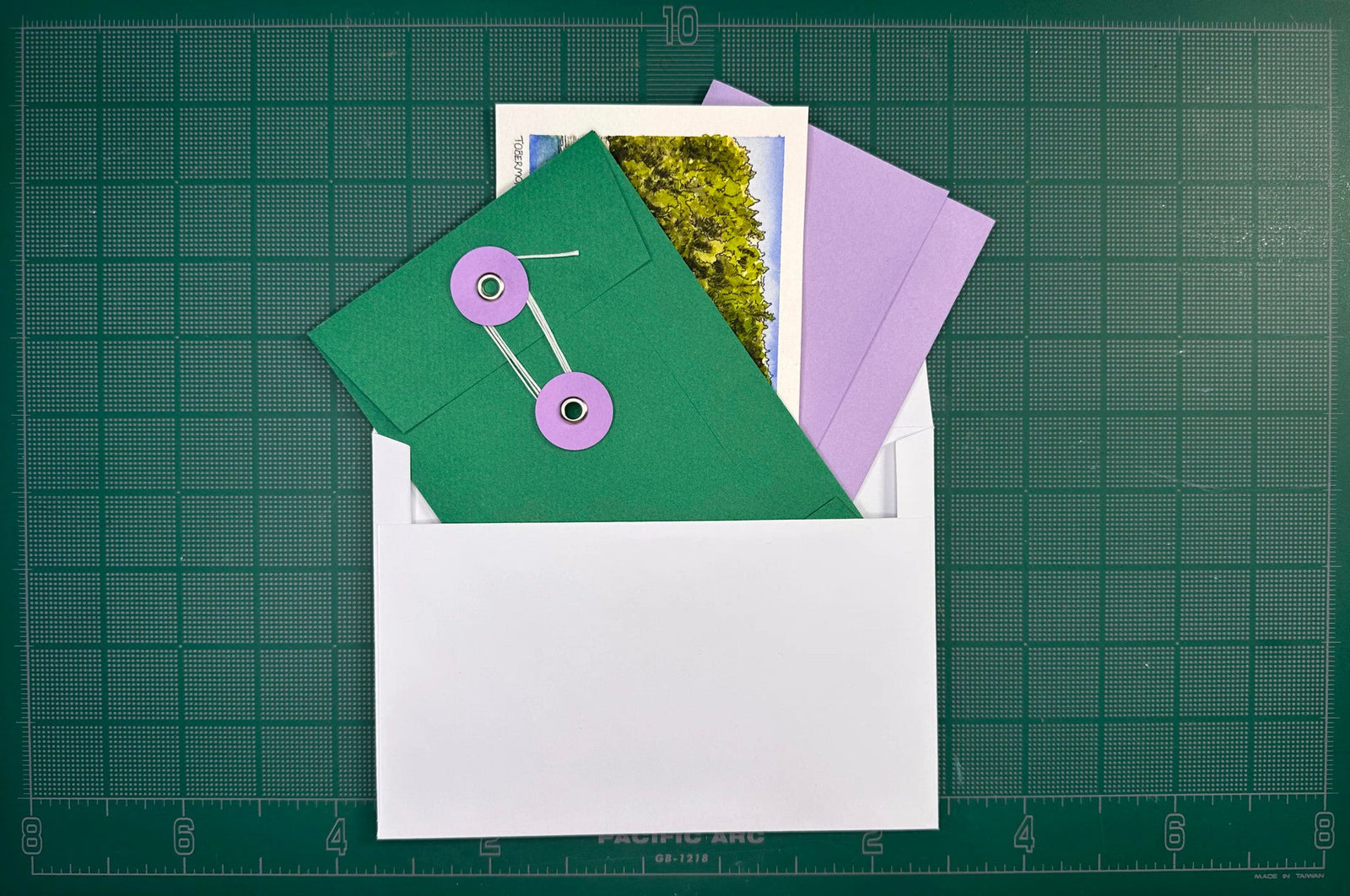 A handmade green envelope tied with string, holding a watercolour print and a letter, tucked into its outer mailing envelope