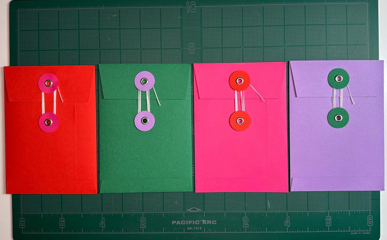 Four handmade eyelet-closure envelopes in red, green, pink and purple, laid out on a green cutting mat
